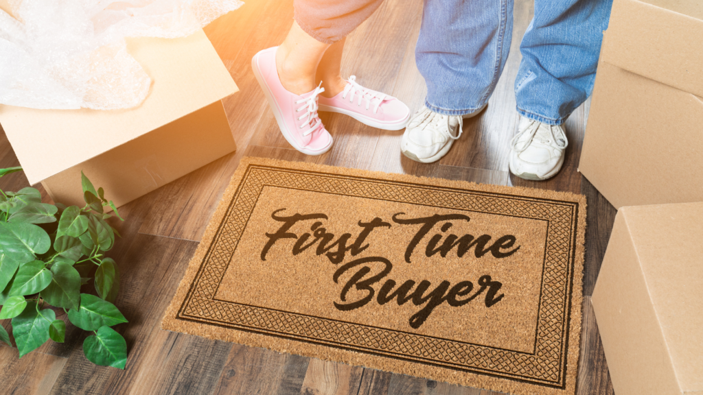 A man and a woman are standing over a doormat that says "first-time buyer" next to moving boxes and plants.