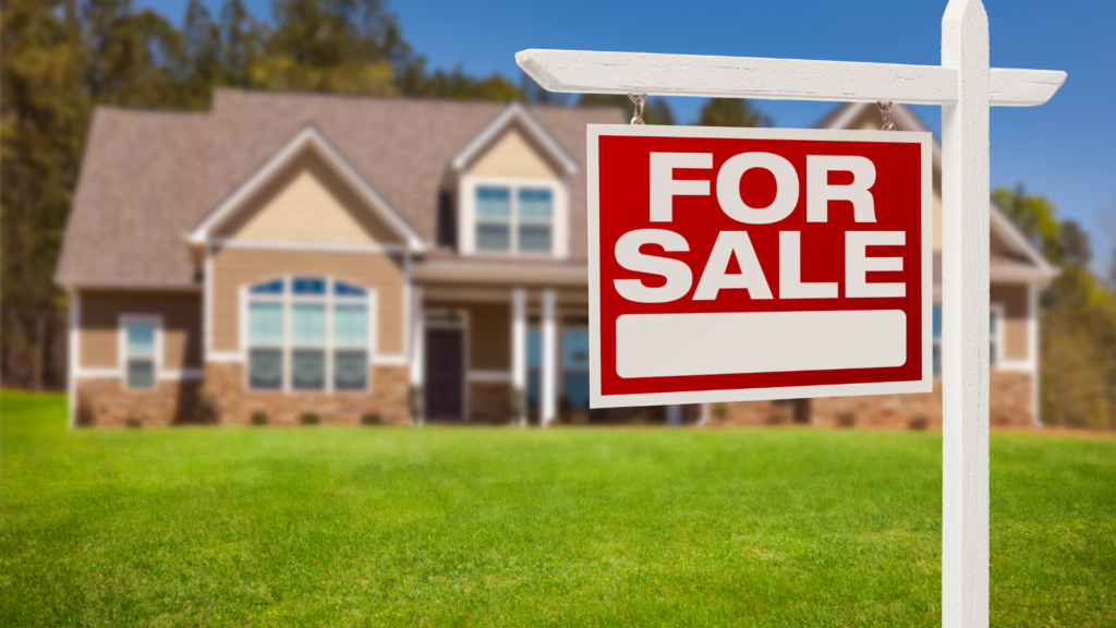 There's a red "for sale" sign outside of a home with blue sky and green grass. 