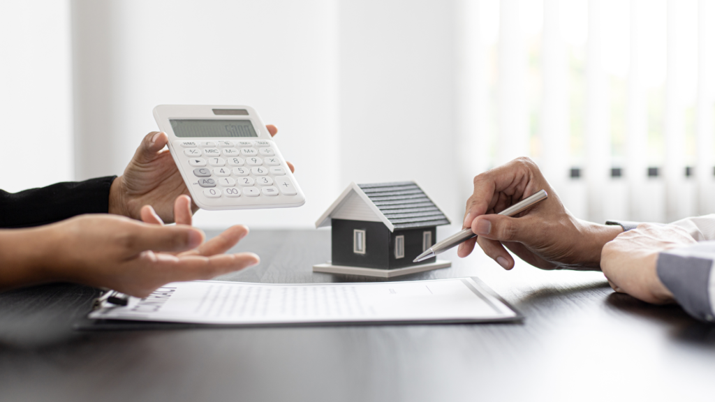 Someone is holding a calculator while another person holds a pen looking over paperwork for purchasing a new assumable mortgage home. 