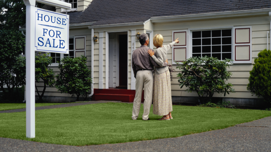 A couple is standing in the front yard of a home with a "house for sale" sign  for a blog titled "FHA Assumable Mortgage Guidelines Everyone Needs to Know."