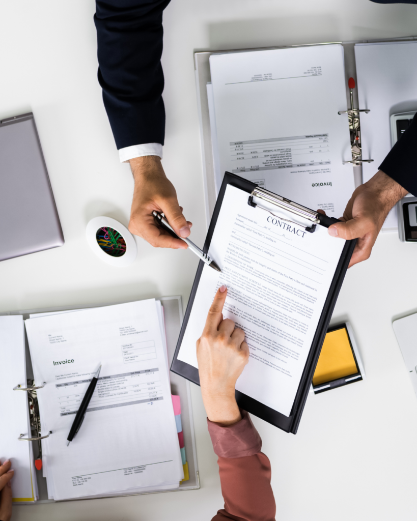 A picture of someone signing a contract. The lawyer is holding a clipboard and pen while the signee points to something on the page.